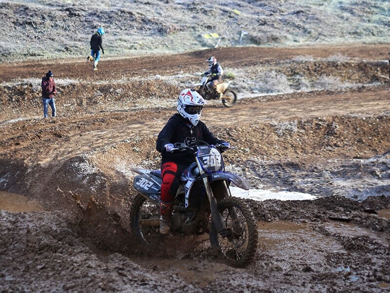 Pilote de motocross en action sur circuit boueux, tenue noire et casque rouge, numéro 331, spectateurs et autres compétiteurs en arrière-plan