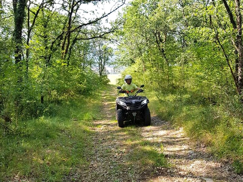 Conducteur de quad traversant un chemin forestier boueux, arbres denses des deux côtés.