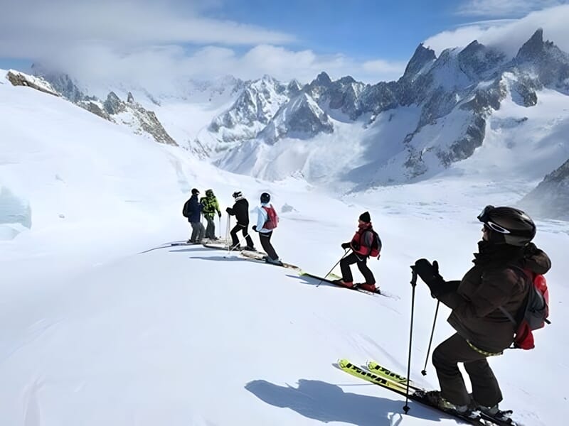 Un groupe de skieurs sur une piste enneigée avec des montagnes escarpées en arrière-plan et un ciel clair.