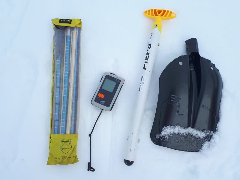 Équipement de sécurité pour avalanche composé d'un appareil de recherche de victime d'avalanche (DVA), d'une sonde et d'une pelle, posés sur la neige.