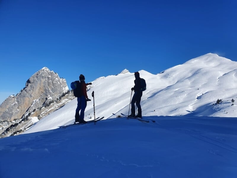 Deux skieurs de randonnée observent la vue sur des sommets enneigés sous un ciel bleu intense.