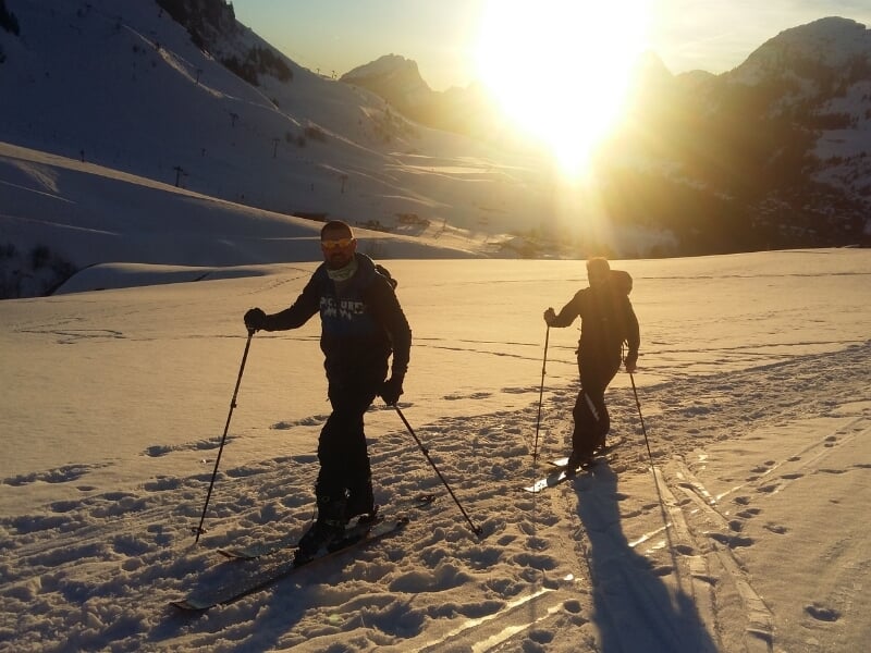 Deux skieurs de randonnée progressent sur une pente neigeuse avec le soleil couchant en arrière-plan, créant une ambiance dorée.