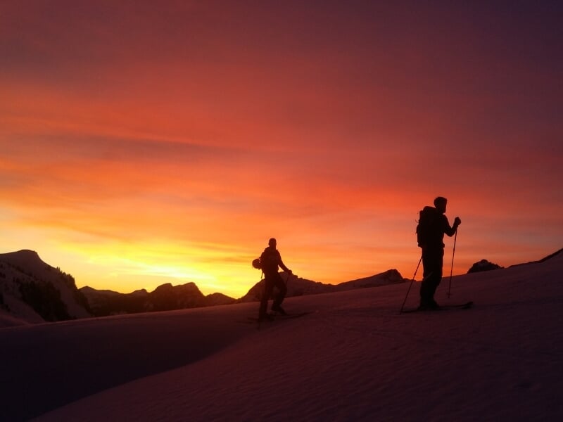 Silhouettes de deux skieurs de randonnée contre un ciel orangé et rose vibrant au coucher du soleil.