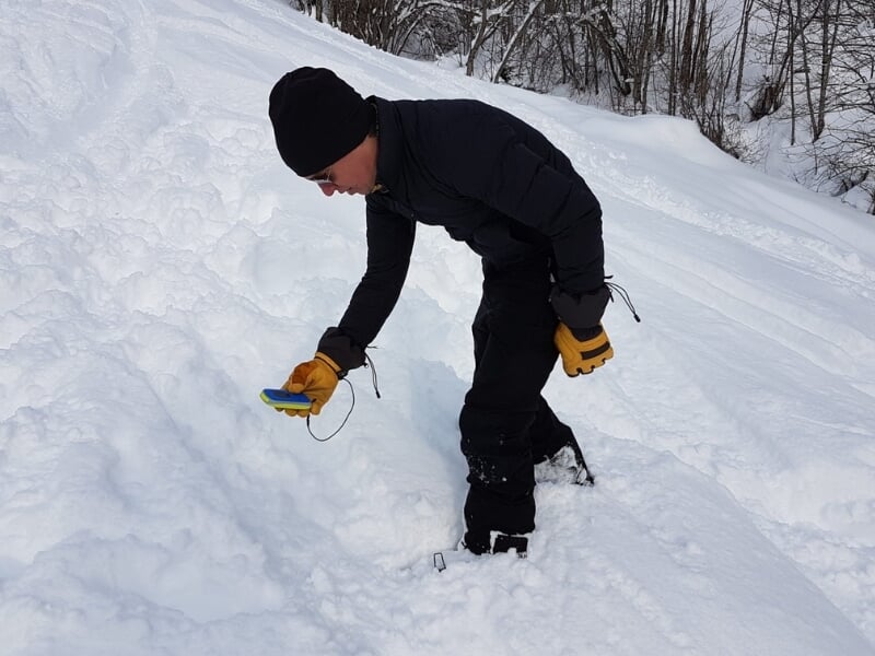 Homme en tenue d'hiver noire utilisant un détecteur de métal jaune et bleu dans une zone enneigée avec des arbres en arrière-plan.