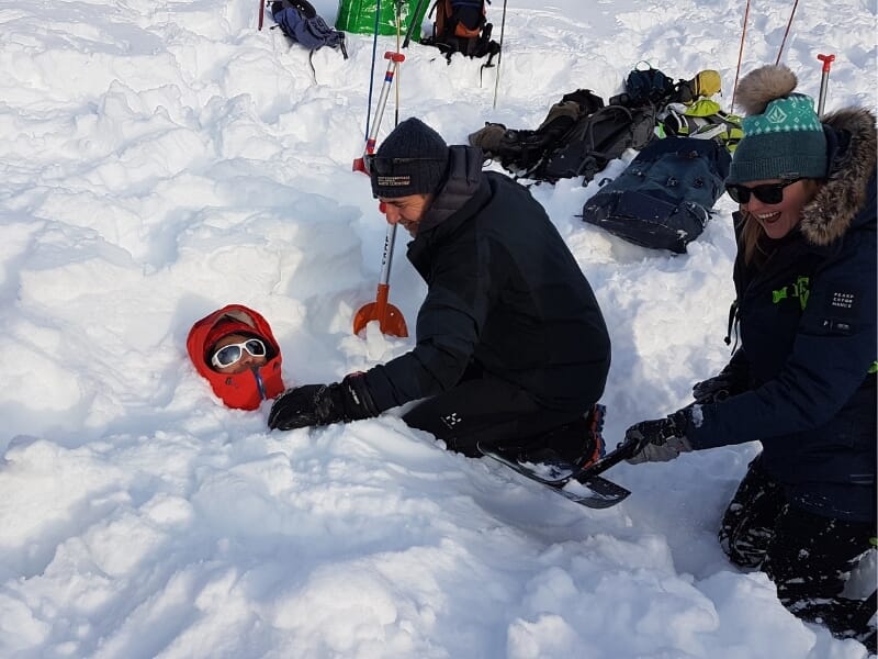 Deux personnes pratiquant un exercice de sauvetage en avalanche, l'une déterrant une tête mannequin rouge avec des lunettes de ski, l'autre observant, avec des équipements dispersés autour dans la nei