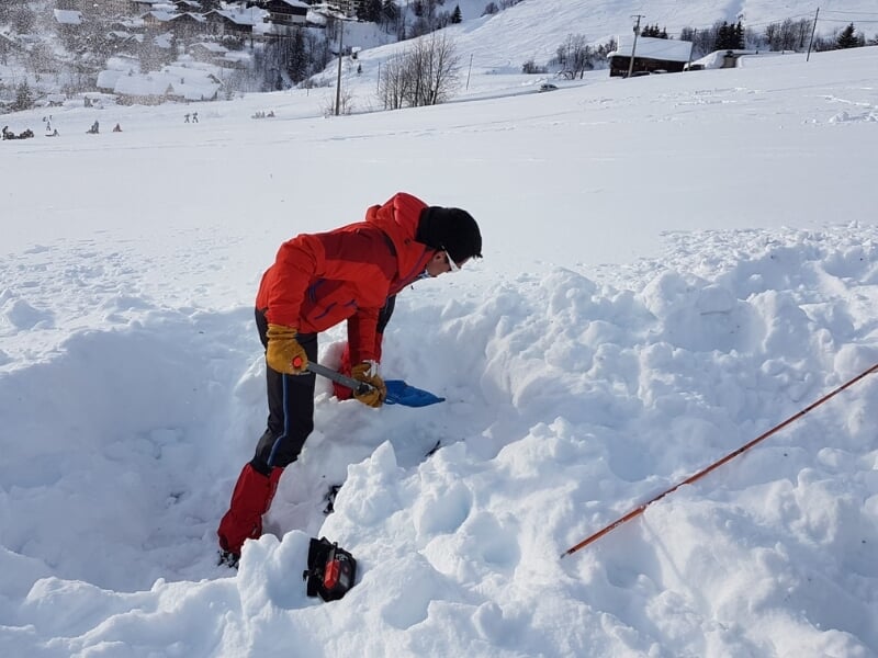 Homme en tenue d'hiver rouge et jaune creusant dans la neige avec un détecteur de métal jaune et bleu et une pelle noire, avec un paysage montagneux en arrière-plan.