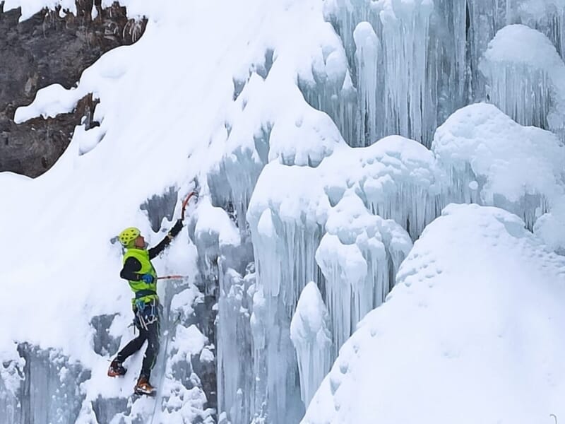 Alpiniste en tenue d'hiver verte et noire, équipé de crampons et piolets, escaladant une cascade de glace verticale avec des formations de glace et de neige autour.