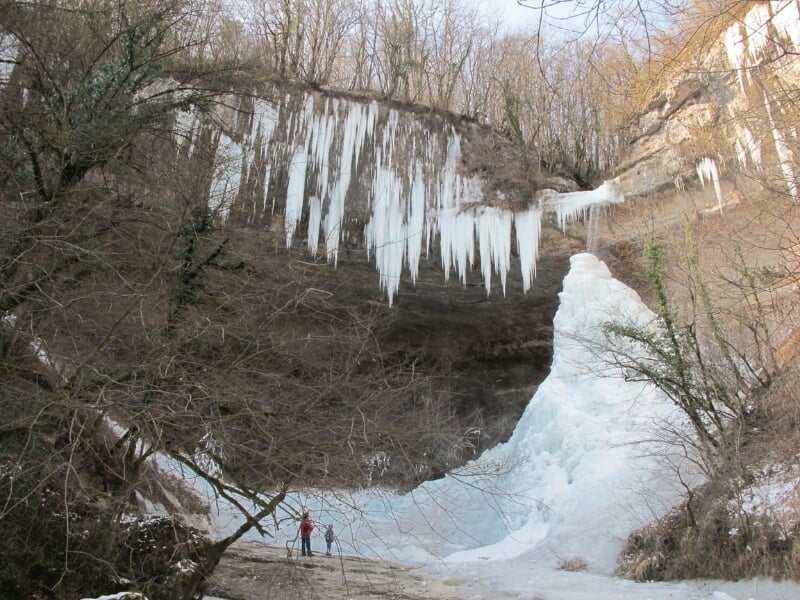 Vue d'une grande cascade de glace avec des stalactites de glace suspendues à une falaise, avec deux petites figures humaines en bas pour l'échelle, illustrant l'immensité de la cascade.