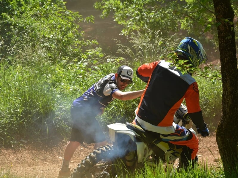 Homme poussant un motard en combinaison orange sur un sentier en forêt, entouré de végétation et de fumée de la moto.