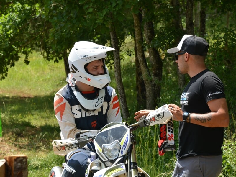 Motard en casque blanc discutant avec un homme en casquette, près d'une moto dans un cadre forestier.