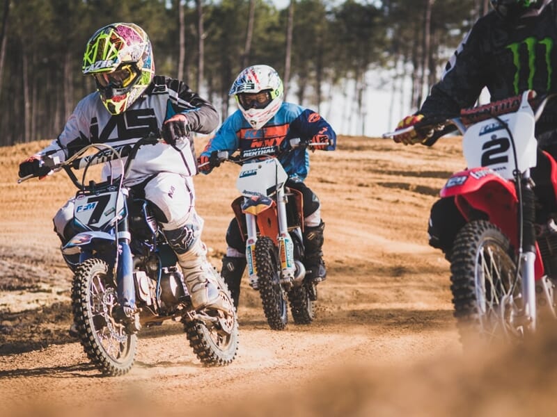 Trois pilotes de motocross sur un circuit de terre, portant des équipements colorés et des casques, en pleine course.