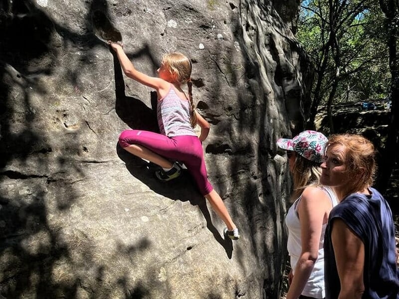 enfant pratiquant l'escalade sur bloc en forêt surveillée par deux adultes