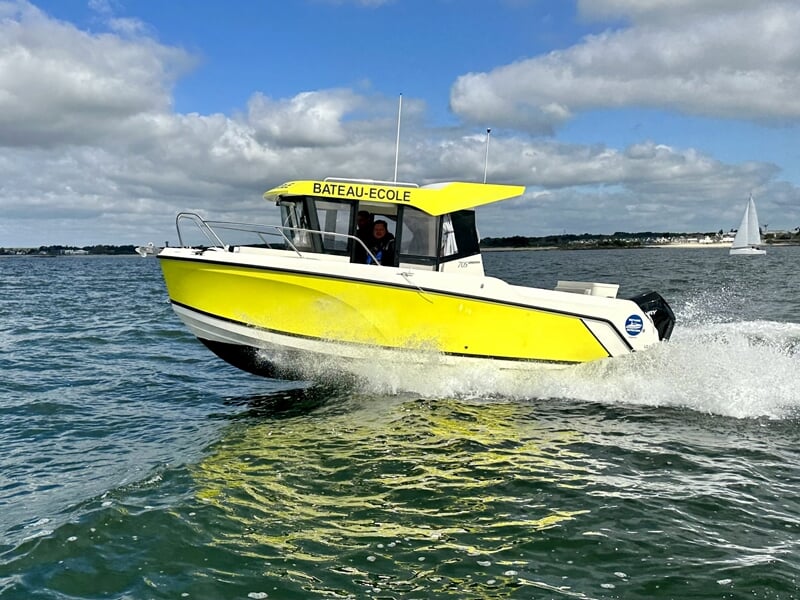 Bateau-école jaune vif naviguant rapidement sur l'eau avec des éclaboussures visibles et d'autres bateaux en arrière-plan, sous un ciel partiellement nuageux
