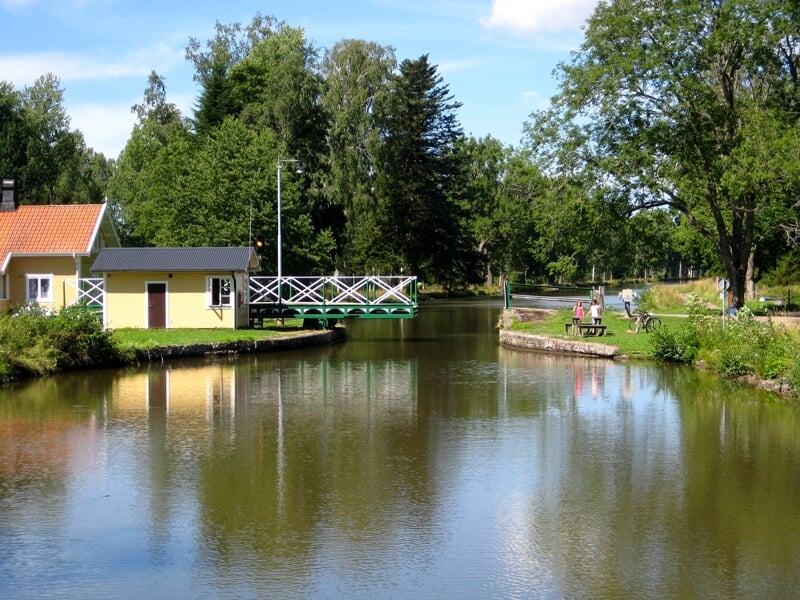 Paisible canal rural avec une petite écluse, une maison jaune, un pont piétonnier, des personnes marchant et un vélo, entouré de verdure sous un ciel bleu avec des nuages légers