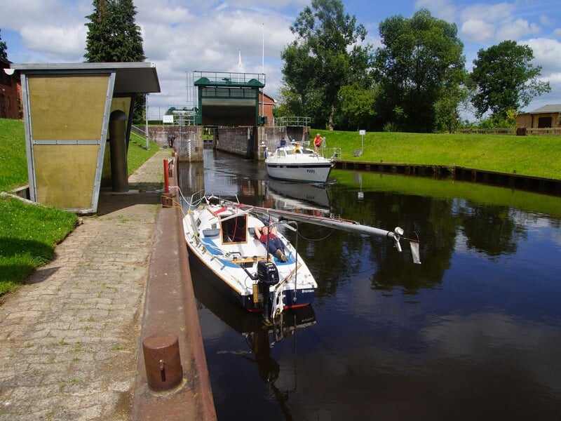 Une écluse avec un petit voilier et un bateau à moteur attendent sur une voie navigable calme, avec des bâtiments et des arbres en arrière-plan sous un ciel nuageux