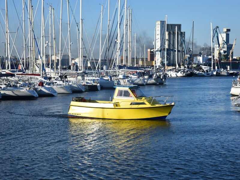 Bateau de plaisance jaune naviguant dans un port de plaisance bondé avec des voiliers ancrés et des grues industrielles en arrière-plan sous un ciel bleu clair