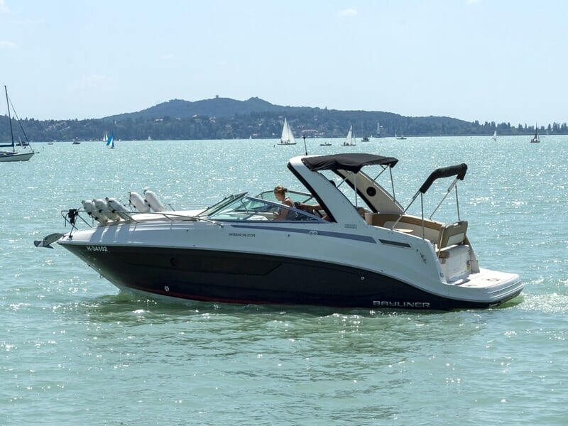 Bateau à moteur de luxe naviguant sur une eau calme avec d'autres voiliers en arrière-plan et une colline verdoyante sous un ciel bleu partiellement nuageux