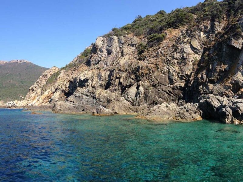Rochers escarpés et végétation méditerranéenne bordant une mer turquoise limpide sous un ciel bleu éclatant.