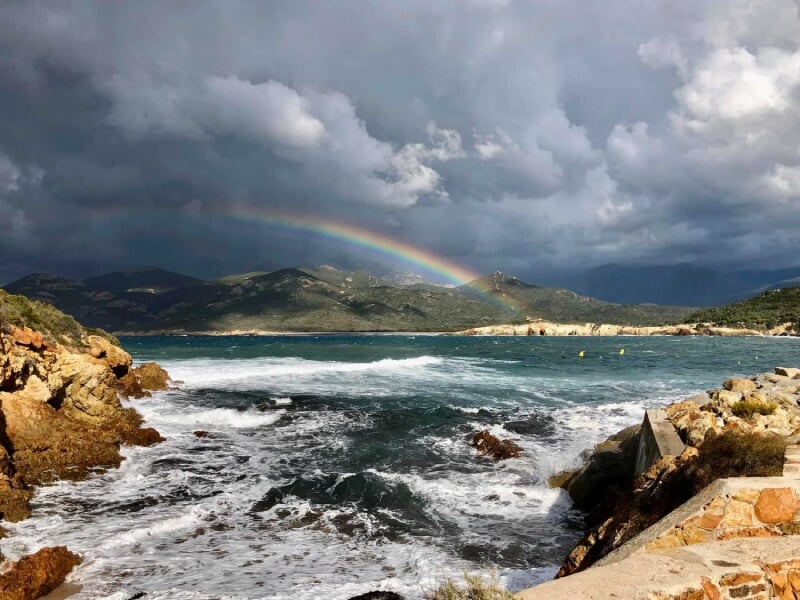 Côte rocheuse avec des vagues déferlantes, un arc-en-ciel vibrant dans le ciel nuageux, montagnes en arrière-plan.