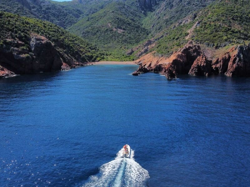 Bateau à moteur voguant sur une mer bleue scintillante, encadré par des montagnes verdoyantes et des falaises rouges.