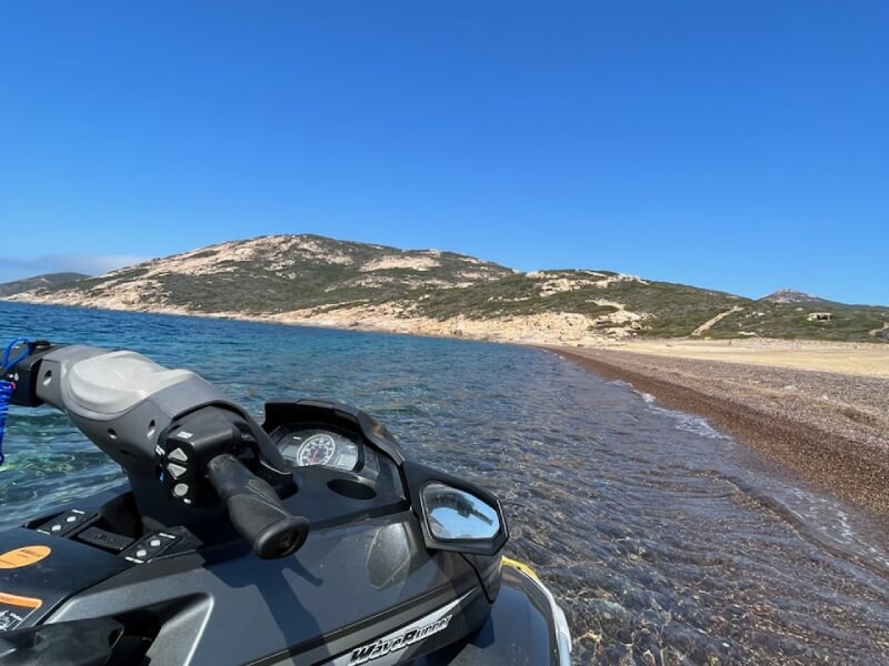 Jet-ski noir amarré près d'une plage de sable avec des collines en arrière-plan, eaux claires et ciel dégagé offrant une vue panoramique.