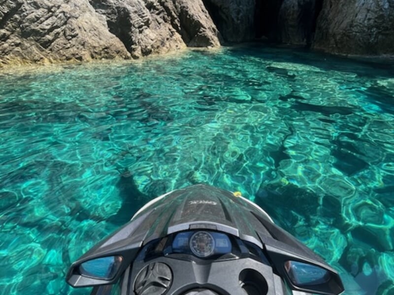 Vue d'un jet-ski noir flottant sur une eau turquoise cristalline entourée de falaises rocheuses, sous un ciel bleu éclatant.