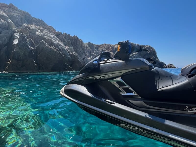 Jet-ski noir naviguant près de falaises rocheuses avec une eau turquoise scintillante, sous un soleil éclatant et un ciel bleu pur.