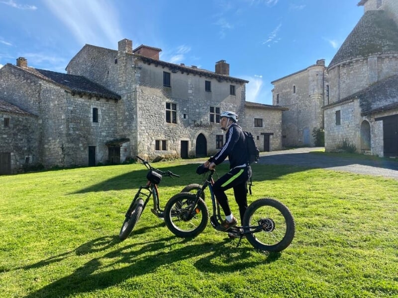 Une personne debout sur une trottinette électrique regarde le paysage, devant une bâtisse en pierre.