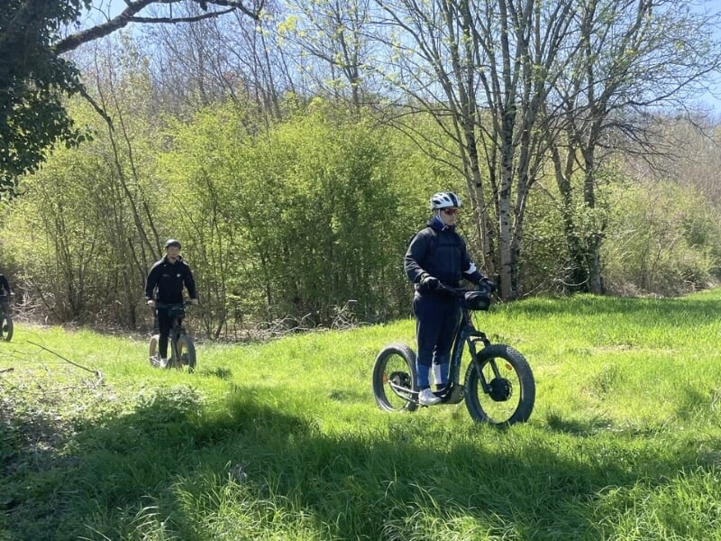 Deux personnes pilotent des trottinettes électriques dans une praire à l'orée d'un bois.