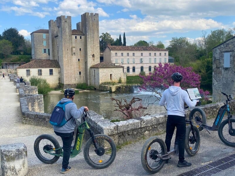 Deux personnes en trottinettes électriques regardent le paysage composé d'une rivière et d'un chateau médiéval.