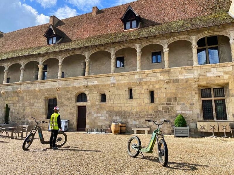 Une personne debout à côté de deux trottinettes électriques admire un bâtiment anciens à voutes.