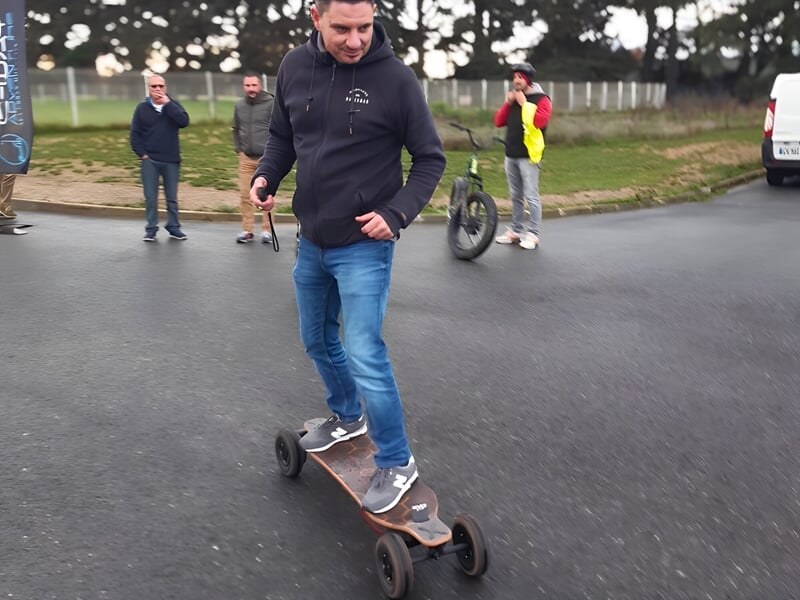 Un homme en sweat noir et jean bleu se tient debout sur un skate électrique sur une route goudronnée.