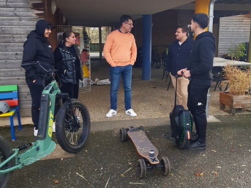 Un groupe de cinq personnes discutent devant l'accueil de location d'engins électriques. Sur le sol, un skate électrique et une trottinette électrique.