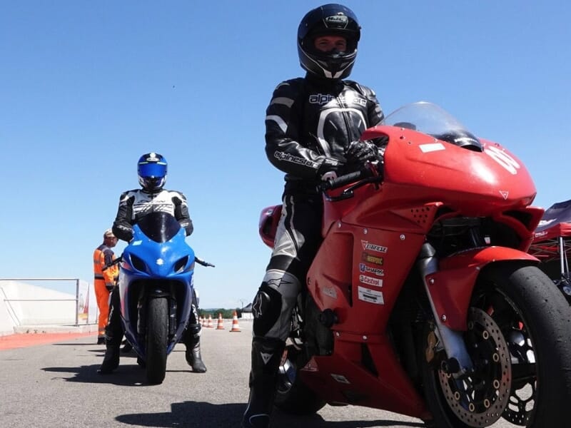 Deux motocyclistes en combinaison de cuir, l'un sur une moto bleue et l'autre sur une moto rouge, se préparant à partir sur une piste avec des cônes de signalisation en arrière-plan.