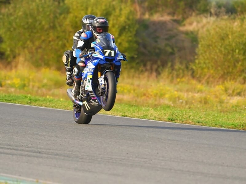 Un motocycliste effectue une roue arrière sur une moto de sport bleue portant le numéro 71 sur une piste de course avec de la végétation en arrière-plan.