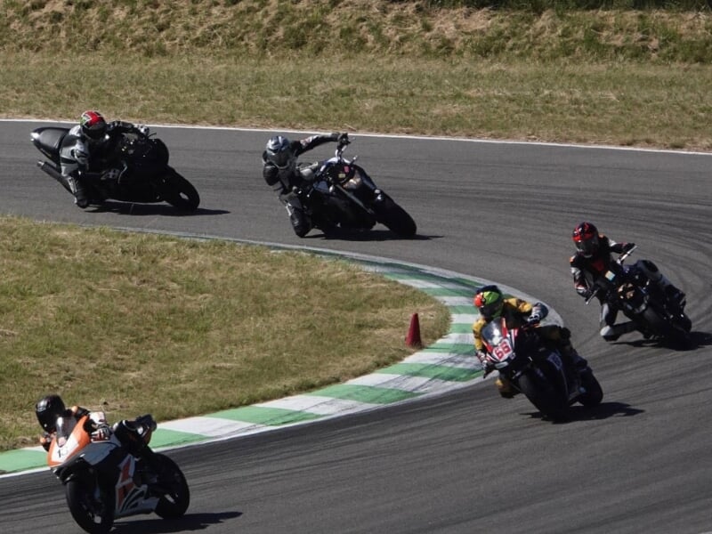 Plusieurs motocyclistes en combinaison de course noire prennent un virage sur une piste de course avec des cônes de signalisation visibles à l'extérieur du virage.