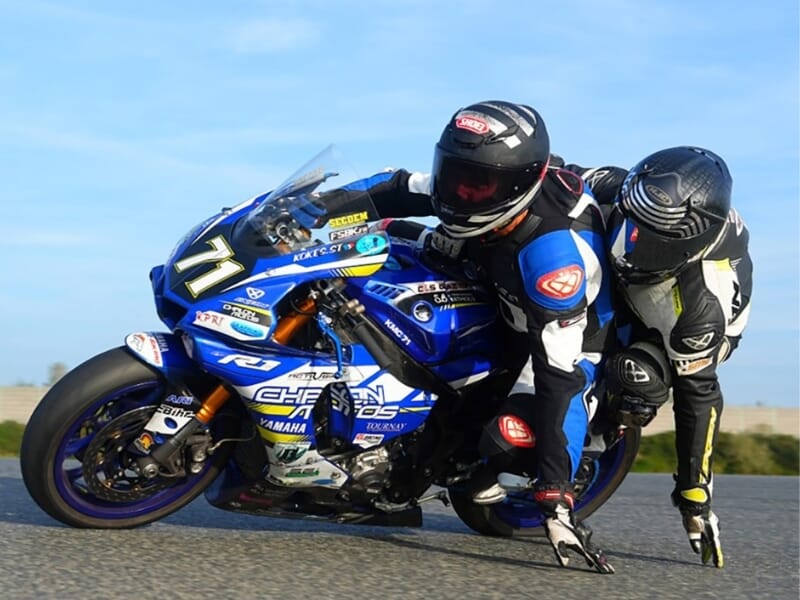 Deux motocyclistes en tandem sur une moto de course bleue, le passager en position de course, sur une piste de course avec le ciel clair en arrière-plan.