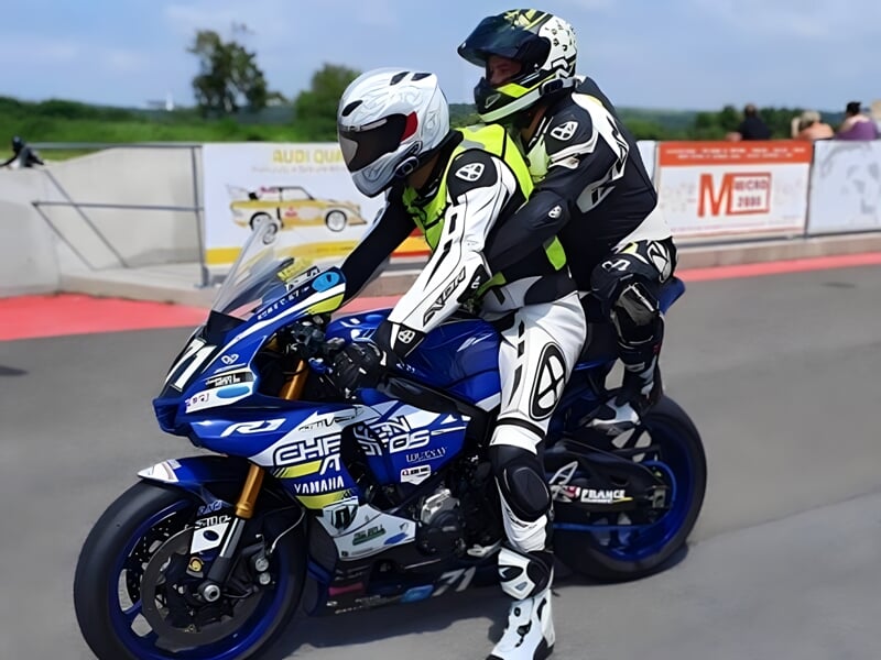 Deux motards en combinaison de course, le passager arrière en combinaison verte et noire et le conducteur en blanc et noir, sur une moto de sport Yamaha bleue et blanche sur une piste de course.