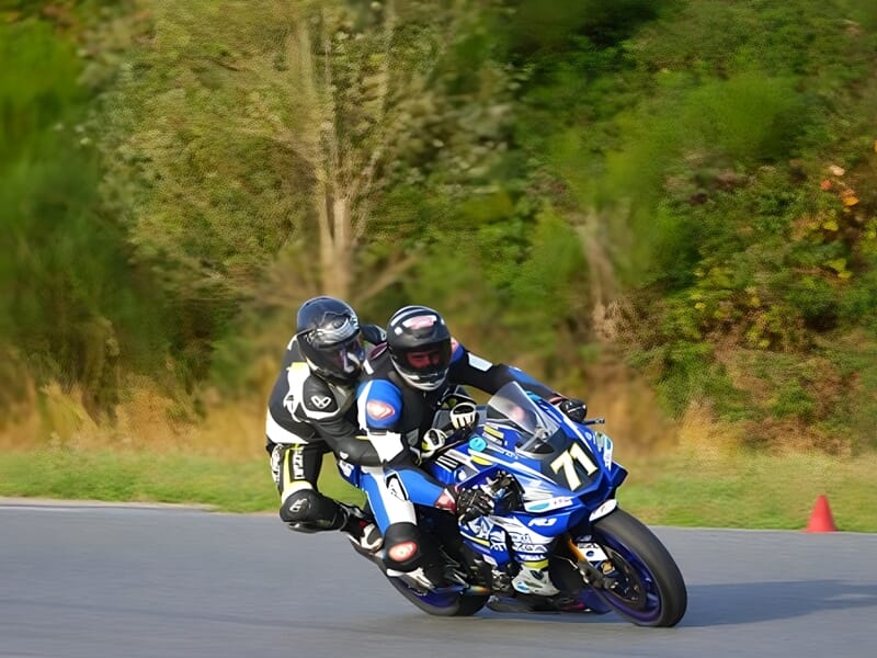 Deux motards en action sur la moto de sport Yamaha, penchés dans un virage sur une piste de course, avec un arrière-plan flou de verdure et d'arbres indiquant une vitesse élevée.