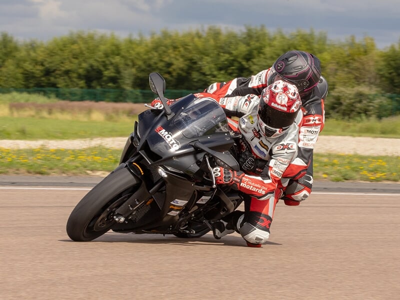 Moto sportive noire dans une courbe serrée avec les pilotes inclinés, entourée de verdure.