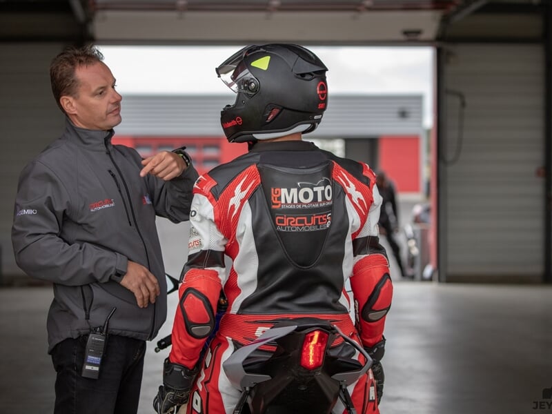 Instructeur en veste grise donnant des consignes à un pilote en tenue rouge et noire, équipé d’un casque intégral, dans un box de circuit automobile