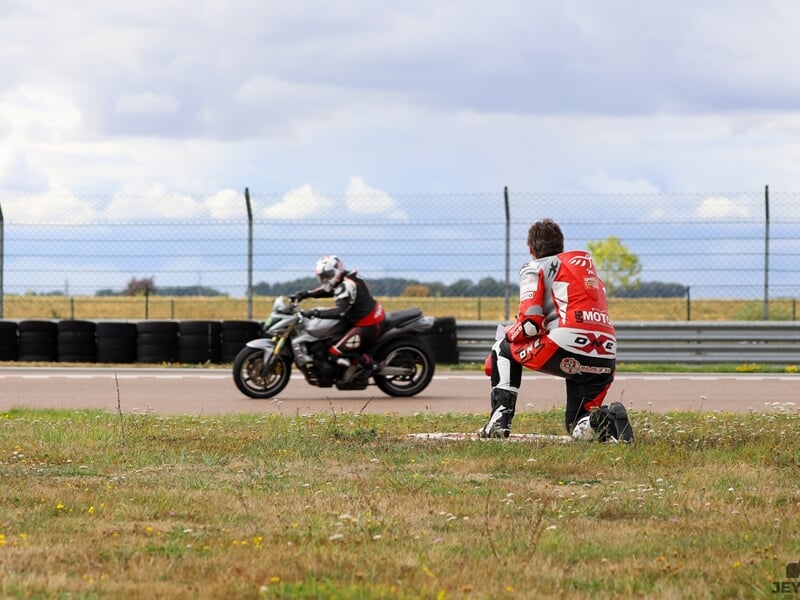 Moniteur en combinaison rouge et blanche observant un motard sur une moto en plein virage, le tout sur un circuit entouré d’herbe et de pneus de sécurité