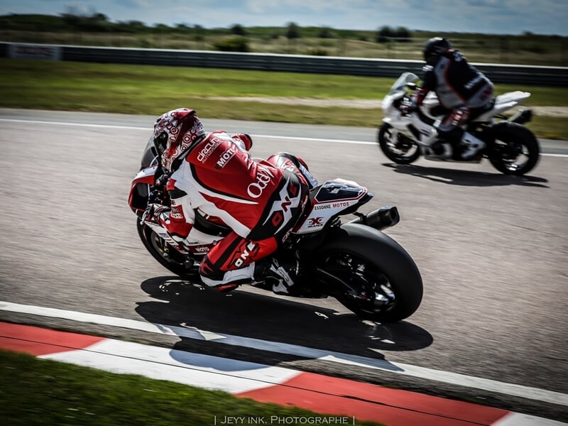 Motard en pleine accélération sur une moto de course rouge et blanche, avec un autre pilote visible en arrière-plan sur une ligne droite de circuit