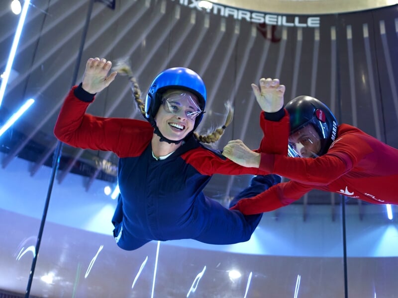 Une femme avec des tresses souriant largement, flotte en chute libre simulée dans un tube vertical, un instructeur en combinaison rouge à sa droite