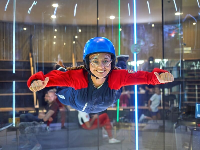 Personne en combinaison de vol et casque bleu souriant à la caméra en position de vol horizontal dans un simulateur de chute libre, avec des spectateurs en arrière-plan