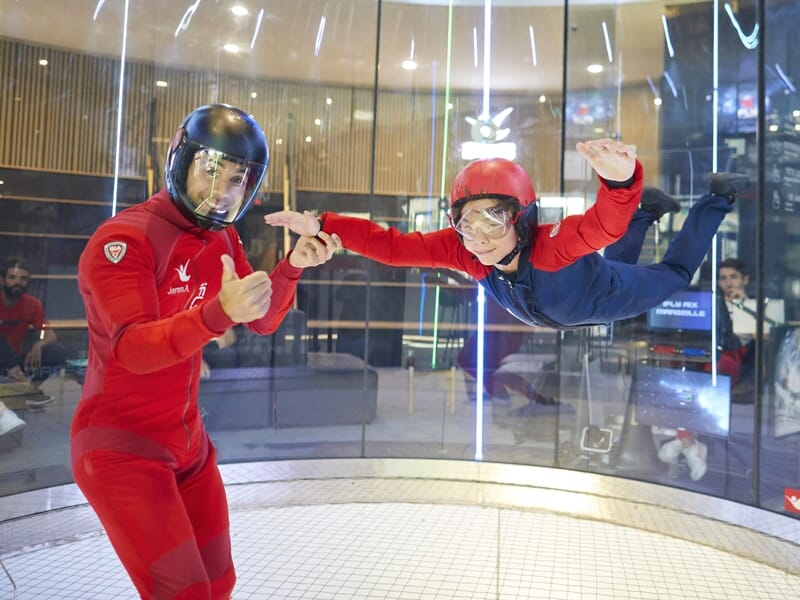 Deux personnes en combinaison de vol, l'une en rouge et l'autre en bleu, pratiquent la chute libre en intérieur avec des gestes de coordination