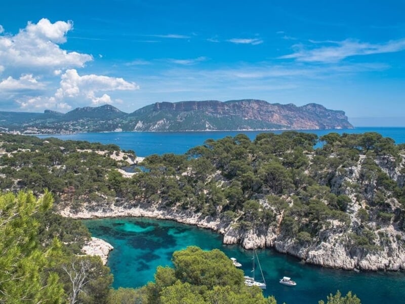 Calanque d'En-Vau avec sa crique turquoise entourée de falaises calcaires et de pins, et des montagnes en arrière-plan sous un ciel bleu