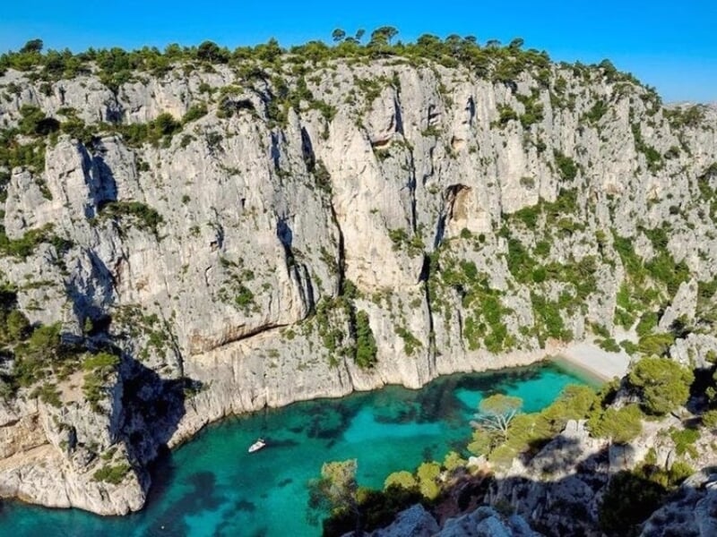 Vue aérienne d'une calanque aux eaux turquoise entourée de falaises rocheuses et de végétation verte dense.
