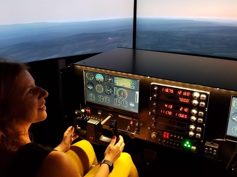 Femme souriante aux commandes d'un simulateur de vol avec plusieurs écrans et instruments de pilotage.