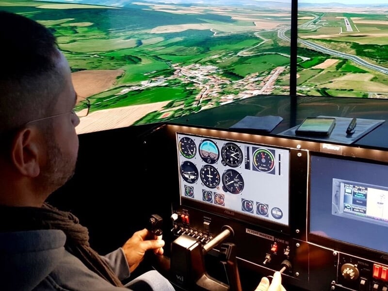Homme concentré pilotant un simulateur d'avion avec vue panoramique sur un paysage rural et instruments de vol.